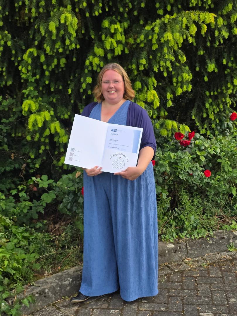 Eine lächelnde Frau in einem blauen Jumpsuit und Cardigan hält ihr Abschlusszertifikat im Freien vor grünen Büschen.