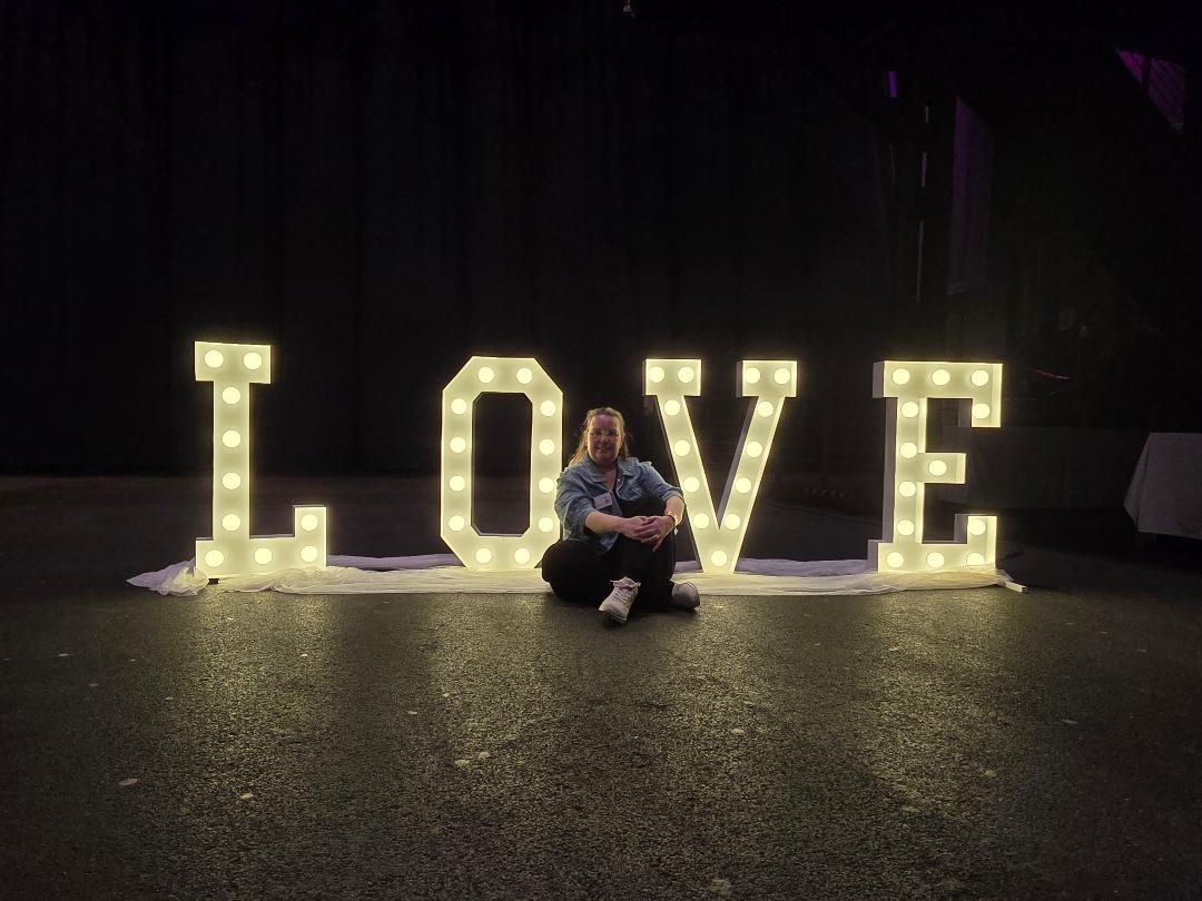 Eine Person sitzt vor großen, beleuchteten LOVE-Buchstaben als Dekoration auf einer dunklen Eventbühne.